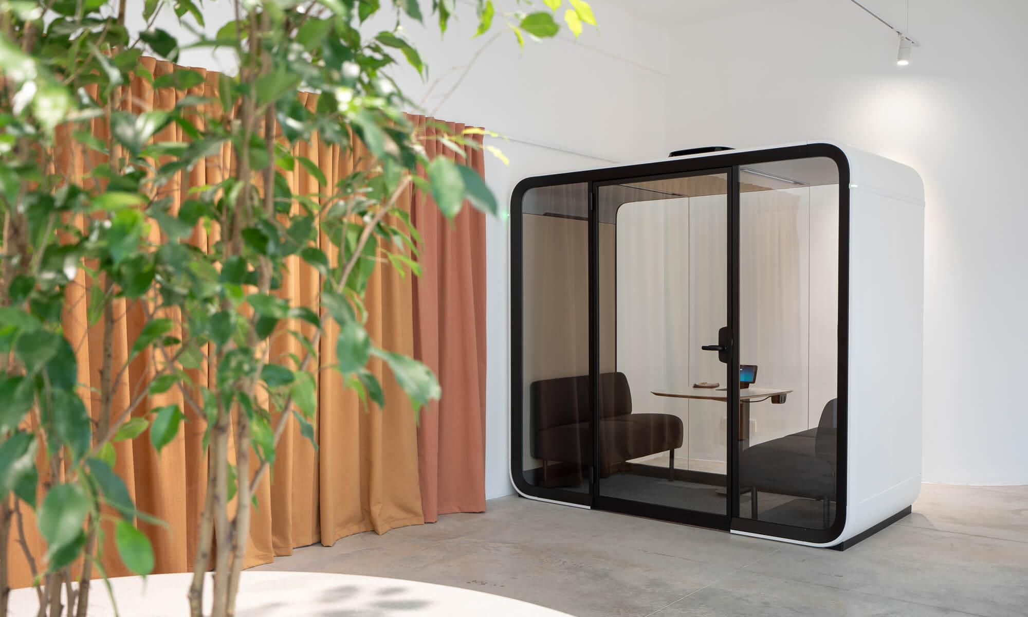 A white Framery Four meeting pod in a space with orange curtains and a plant.