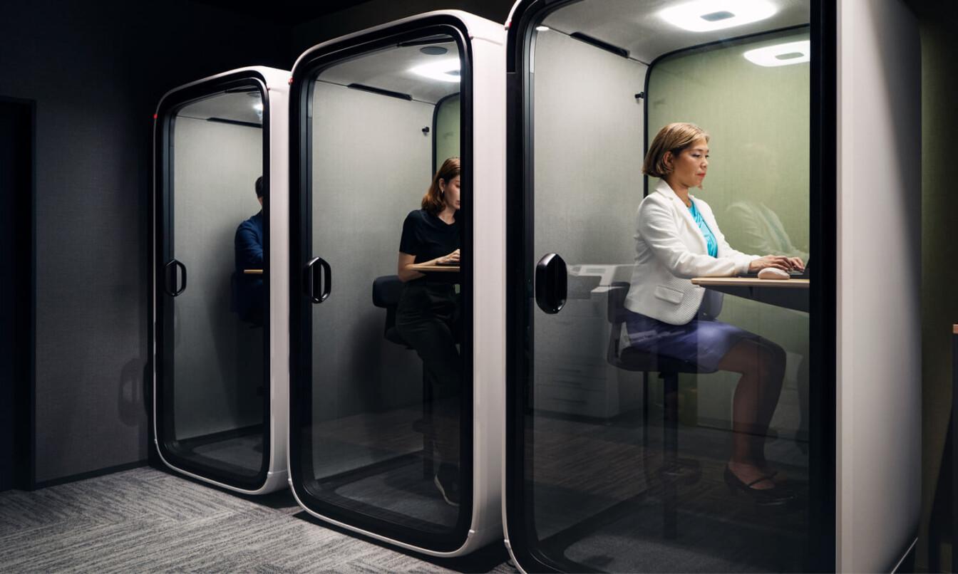 Three people work in modern, glass-enclosed pods, focusing on their tasks. The office environment appears quiet and private, enhancing concentration.