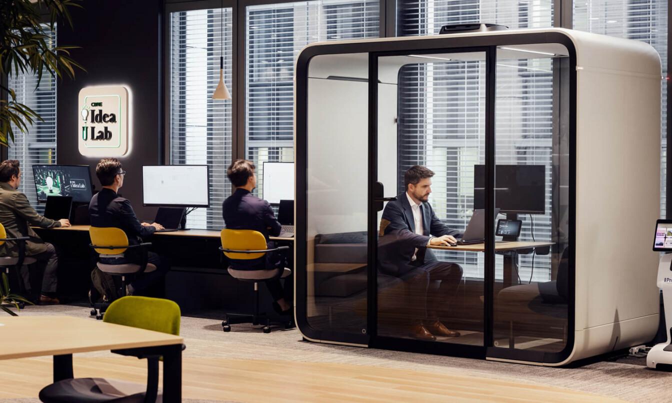 A modern office with people working at desks and a person inside a Framery Four meeting pod, focusing on a laptop. "Idea Lab" sign visible.