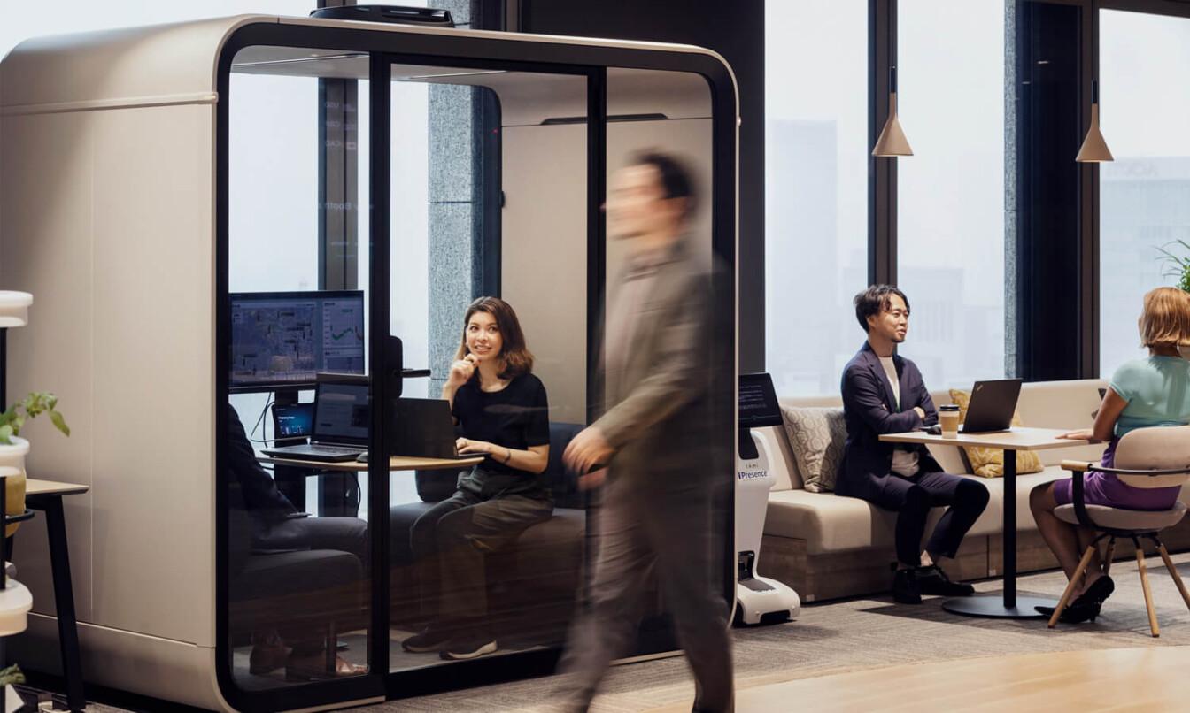 Modern office space with a Framery Four smart meeting pod, people working on laptops, and a person walking by. Bright, well-lit environment.