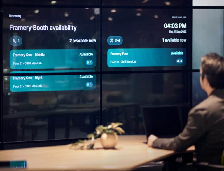 A person sits at a desk viewing digital screen displaying Framery pod availability information, showing time and date. Plant decorates the table.