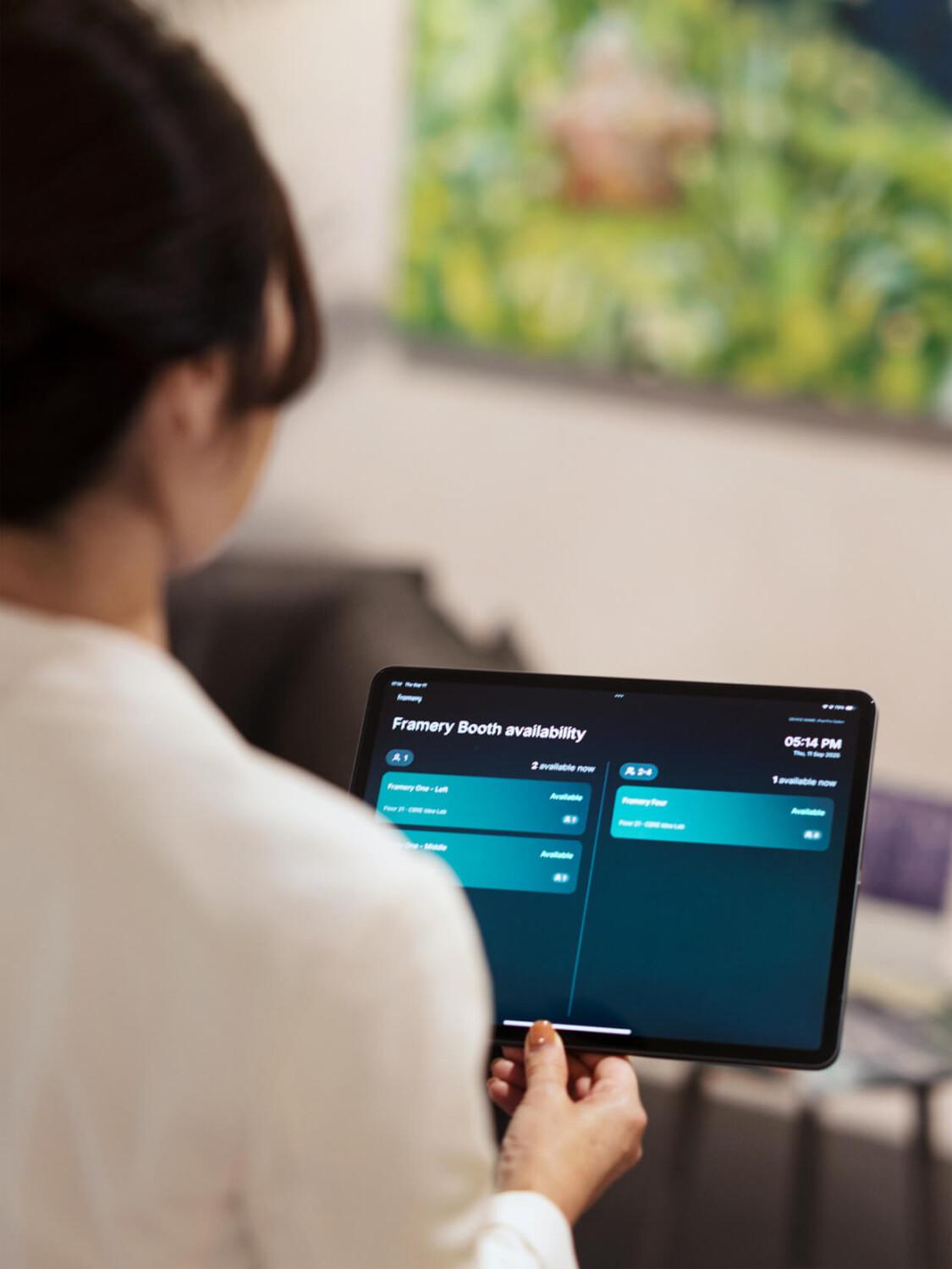 A person is holding a tablet displaying Framery Space Overview in an indoor setting with blurred background artwork.