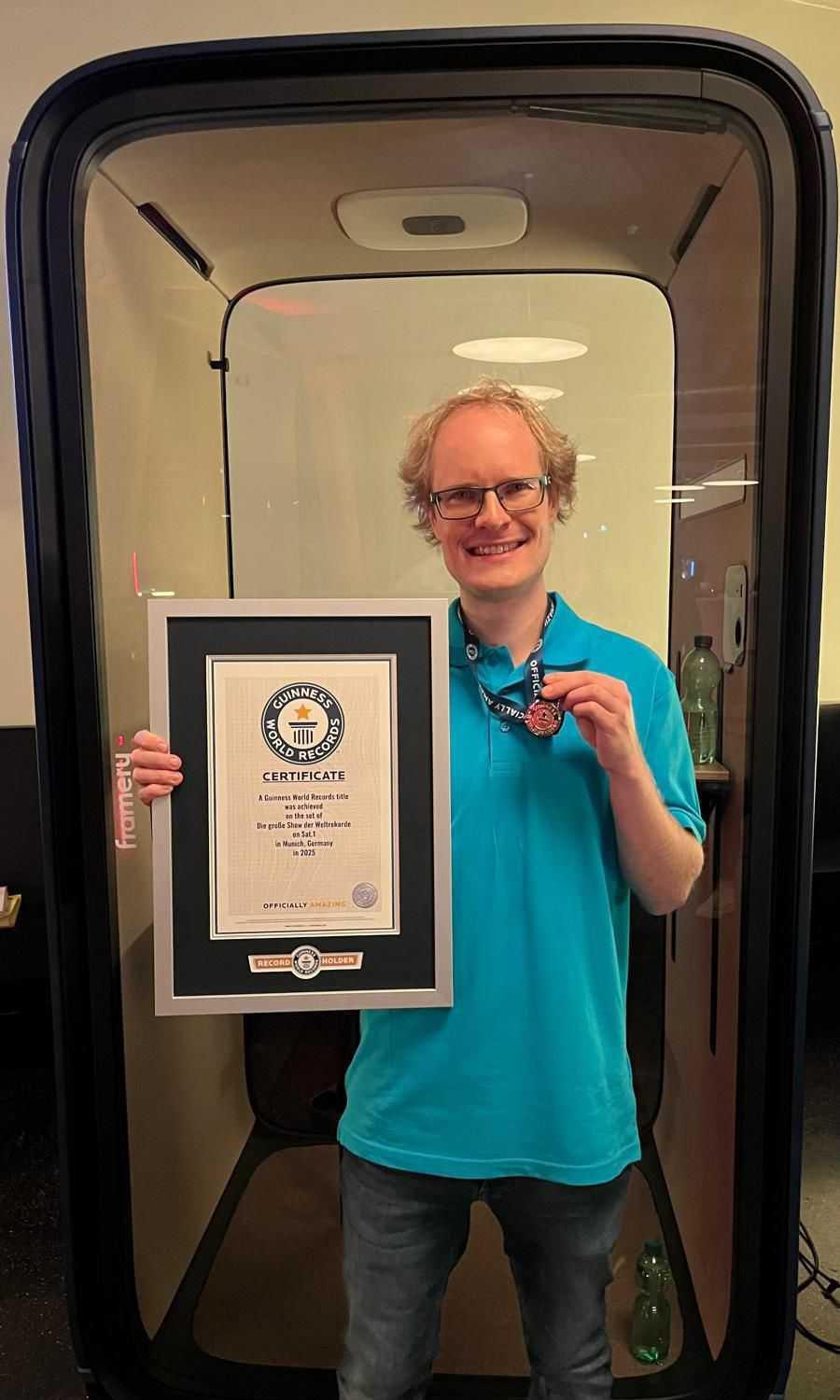 A person in a blue shirt holds a Guinness World Records certificate and medal, standing in front of a Framery soundproof booth.