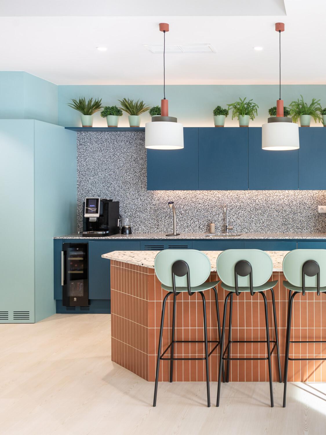 Modern kitchen with teal cabinets, terrazzo backsplash, brown-tiled island, three mint stools, hanging lamps, and potted plants. Bright, minimalist design.