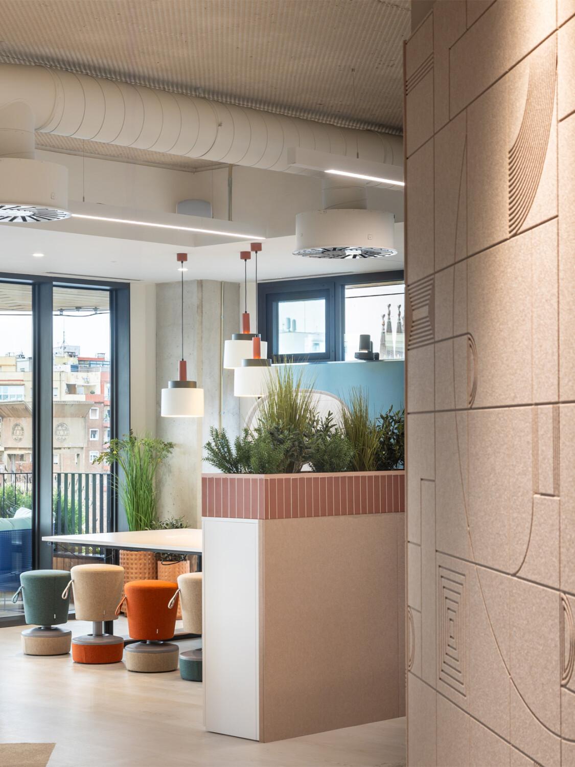 Modern office space with colorful seating, green plants, and large windows. Urban skyline visible outside. Decorative wall design in the foreground.