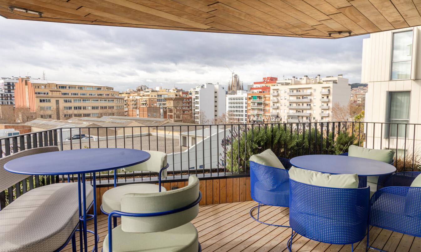 Modern balcony with blue furniture overlooks cityscape of Barcelona. Sagrada Família visible among buildings under cloudy sky. Urban and architectural contrast.
