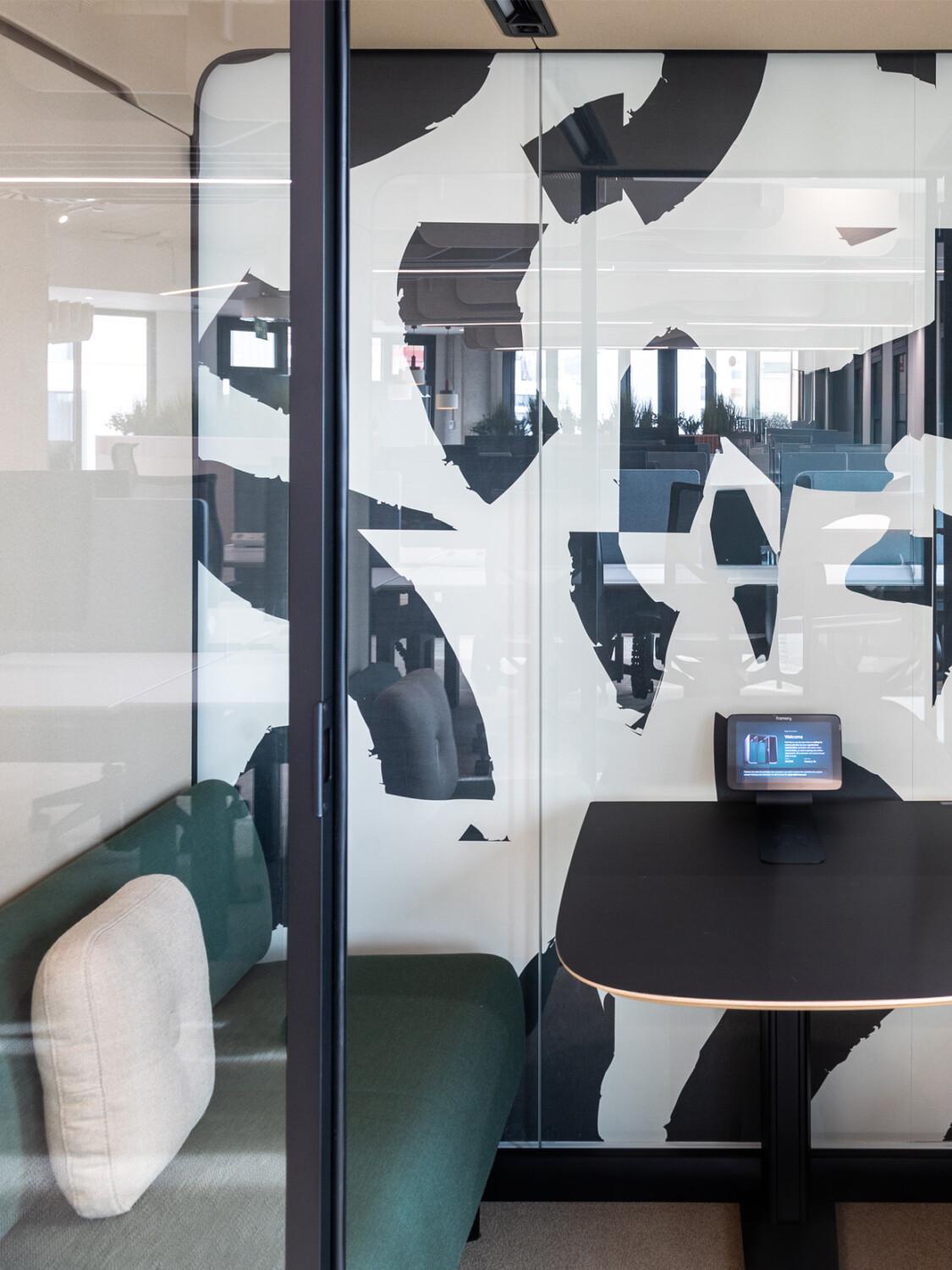 A modern Framery office pod with abstract black-and-white glass art, featuring a small table, bench, and pod's touchscreen panel on table.