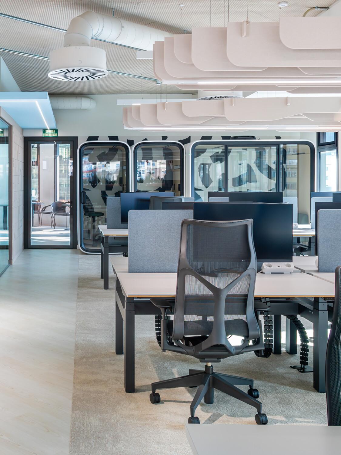 Modern office interior with ergonomic chairs, desks, and bright lighting. Framery smart pods at the back. Open-plan layout with glass walls and well-organized workspaces. No people present.
