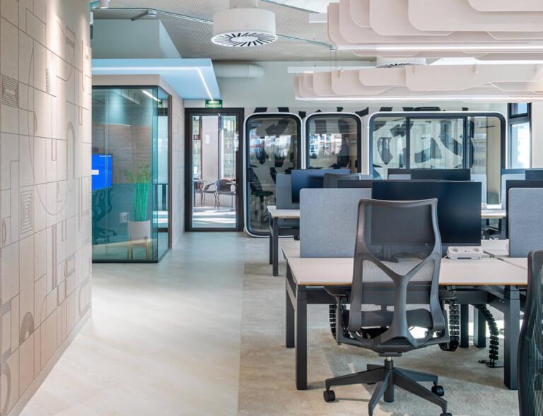 Modern office space with ergonomic chairs, open workstations, glass-walled Framery meeting rooms, and decorative wall designs, creating a bright and collaborative work environment.