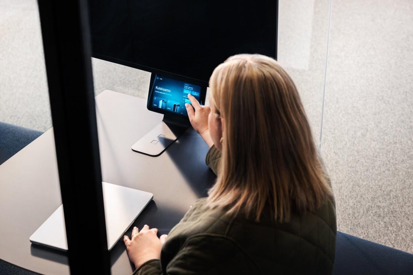 A person interacts with a touch screen at a desk, next to a laptop, in a modern office environment.