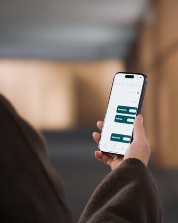 A person holds a smartphone displaying Framery app with meeting spaces listed. Background is blurred, featuring neutral tones, suggesting an indoor environment.