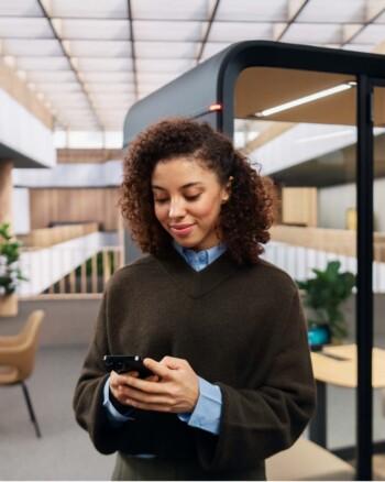 A person with curly hair is using a smartphone in a modern, stylish office environment with glass partitions and natural lighting.