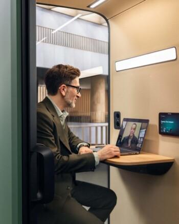 A person in a private booth video conferencing on a laptop, featuring modern interior design with bright lighting and electronic devices visible.