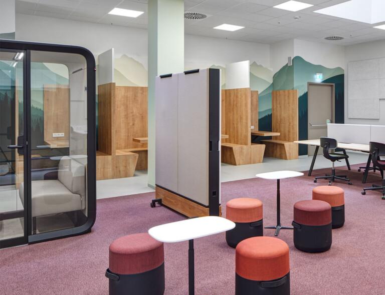 A modern and flexible learning space features partitioned workstations, a private Framery learning pod, colorful stools, and a mountain mural, creating a vibrant and collaborative environment.