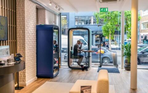 Bright modern lobby with a blue curved phone booth, a glass-walled Framery phone booth with a person inside, and a beige sofa near the entrance.