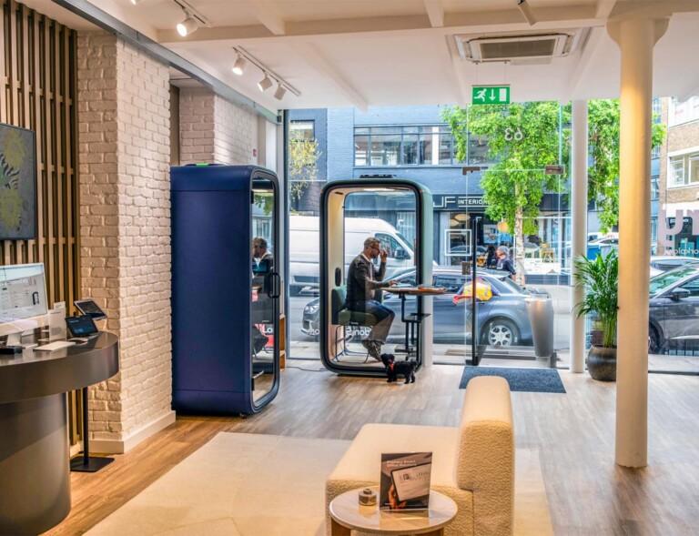 Bright modern lobby with a blue curved phone booth, a glass-walled Framery phone booth with a person inside, and a beige sofa near the entrance.