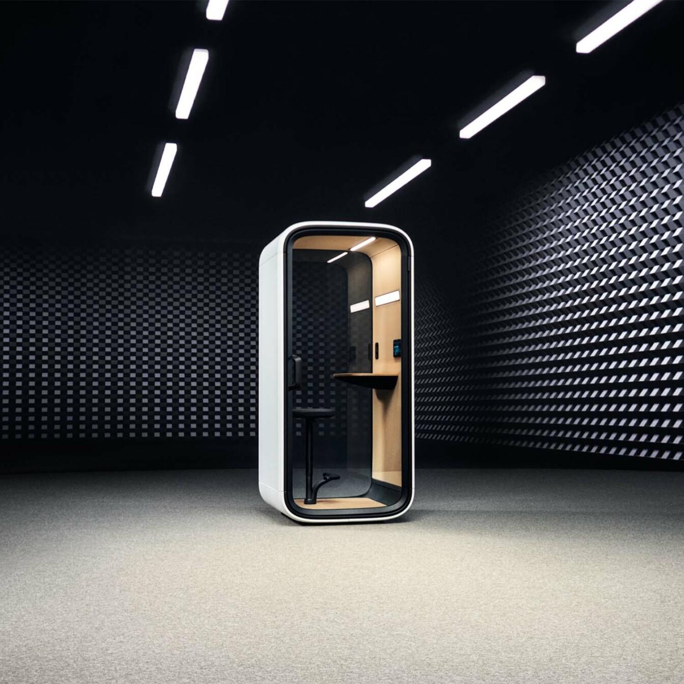 Framery One Compact, a smart office phone booth in an empty room with dark walls and bright lights on the ceiling.