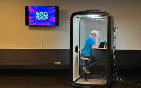 A person inside a soundproof booth works on a laptop. Nearby, a screen displays "Guinness World Records" in a brightly colored graphic.