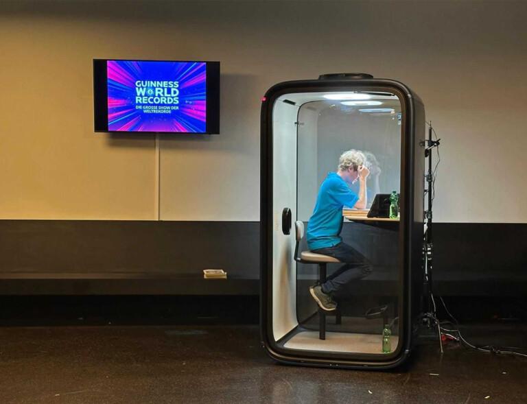 A person inside a soundproof booth works on a laptop. Nearby, a screen displays "Guinness World Records" in a brightly colored graphic.