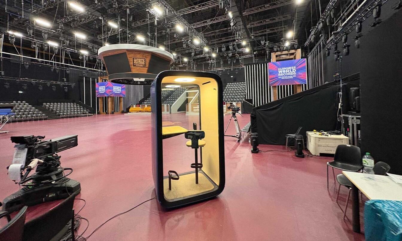 Empty studio with cameras and seating, featuring a phone booth at center. Screens displaying “Guinness World Records” in the background.