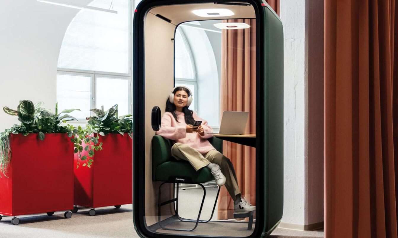 A person sits in a learning pod, wearing headphones, with a laptop on the desk, surrounded by plants.