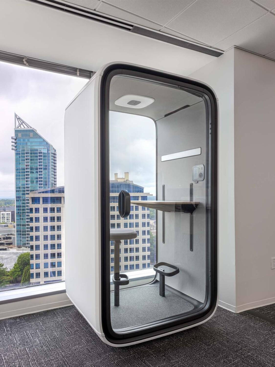 A modern office space contains a sleek, compact phone booth overlooking a cityscape with high-rise buildings and large windows.