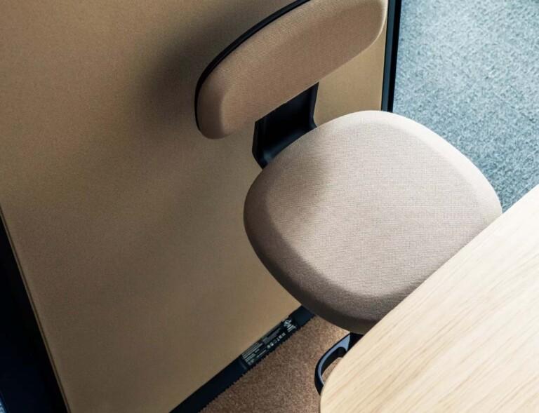 A beige cushioned chair faces a wooden desk, positioned on a carpeted floor, creating a minimalist and modern workspace setting.