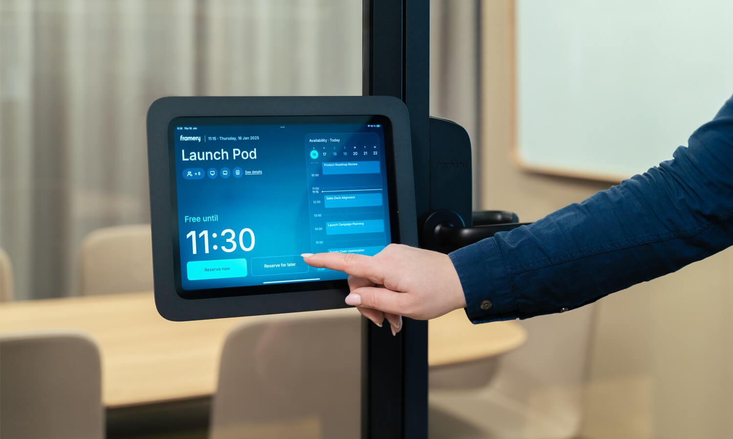 A person interacts with a digital booking screen for room reservation, displaying a modern office environment in the background.