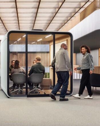 People sit in a modern Framery office pod, conversing. Two individuals walk outside, with contemporary architecture in the background, under a geometric ceiling.