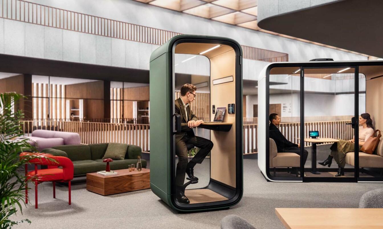 A person having a video call inside a Framery office phone booth with two others having an in-person meeting inside another office pod.