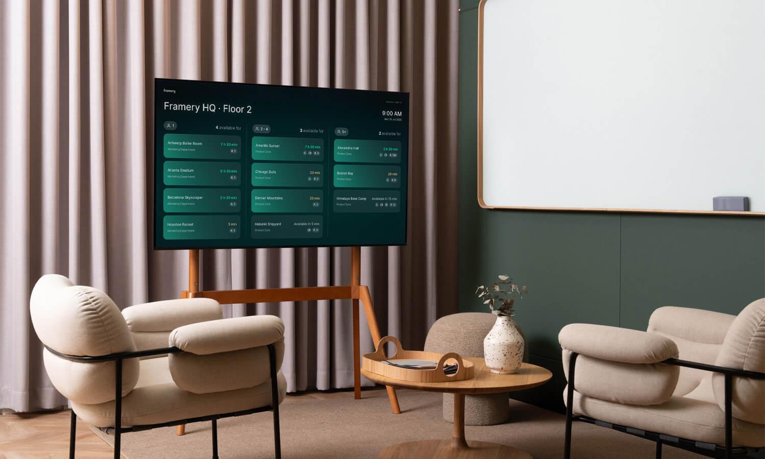 Modern office lounge with neutral chairs, wooden table, and a digital screen displaying room availability at Framery HQ. Whiteboard on the wall.