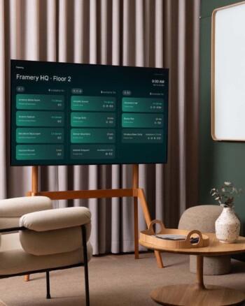 A modern office space with a digital screen displaying Framery Space Overview with meeting room schedules, featuring minimalist furniture and a decorative vase on a round table.