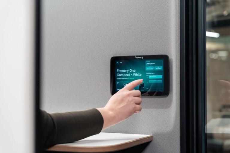 A person interacts with a Framery One Compact touch screen, adjusting reservation settings in a modern office environment.
