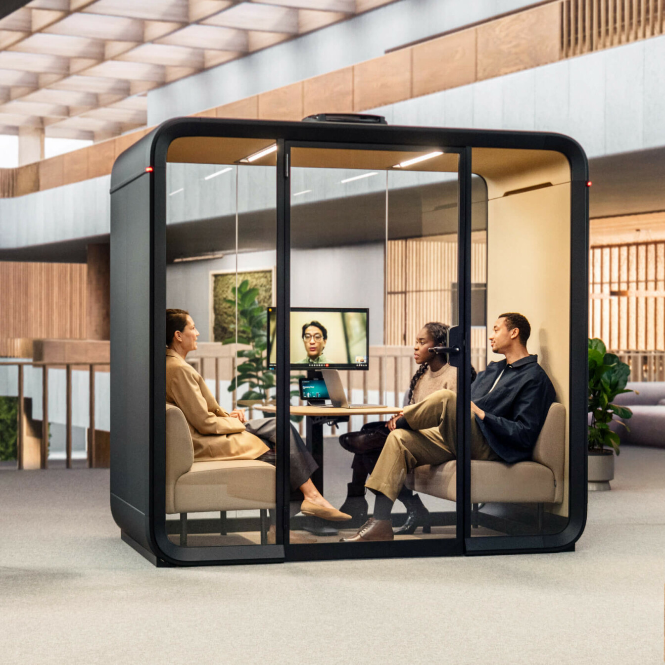Three people seated in a meeting with another person attending through a video call inside a meeting pod in a lounge area of an office. 