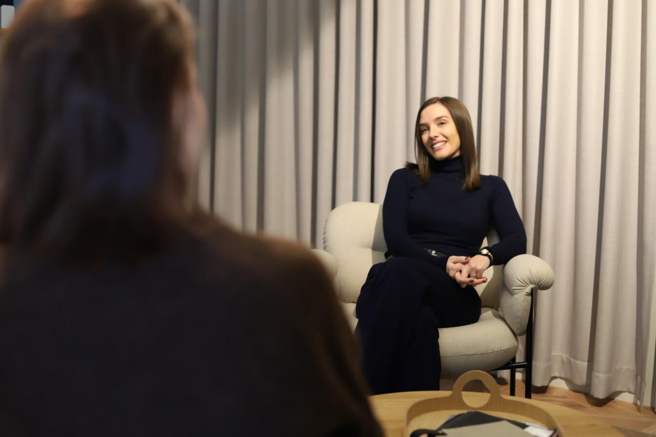 Two people are talking in a room with gray curtains. One person is seated, smiling, on a white chair.