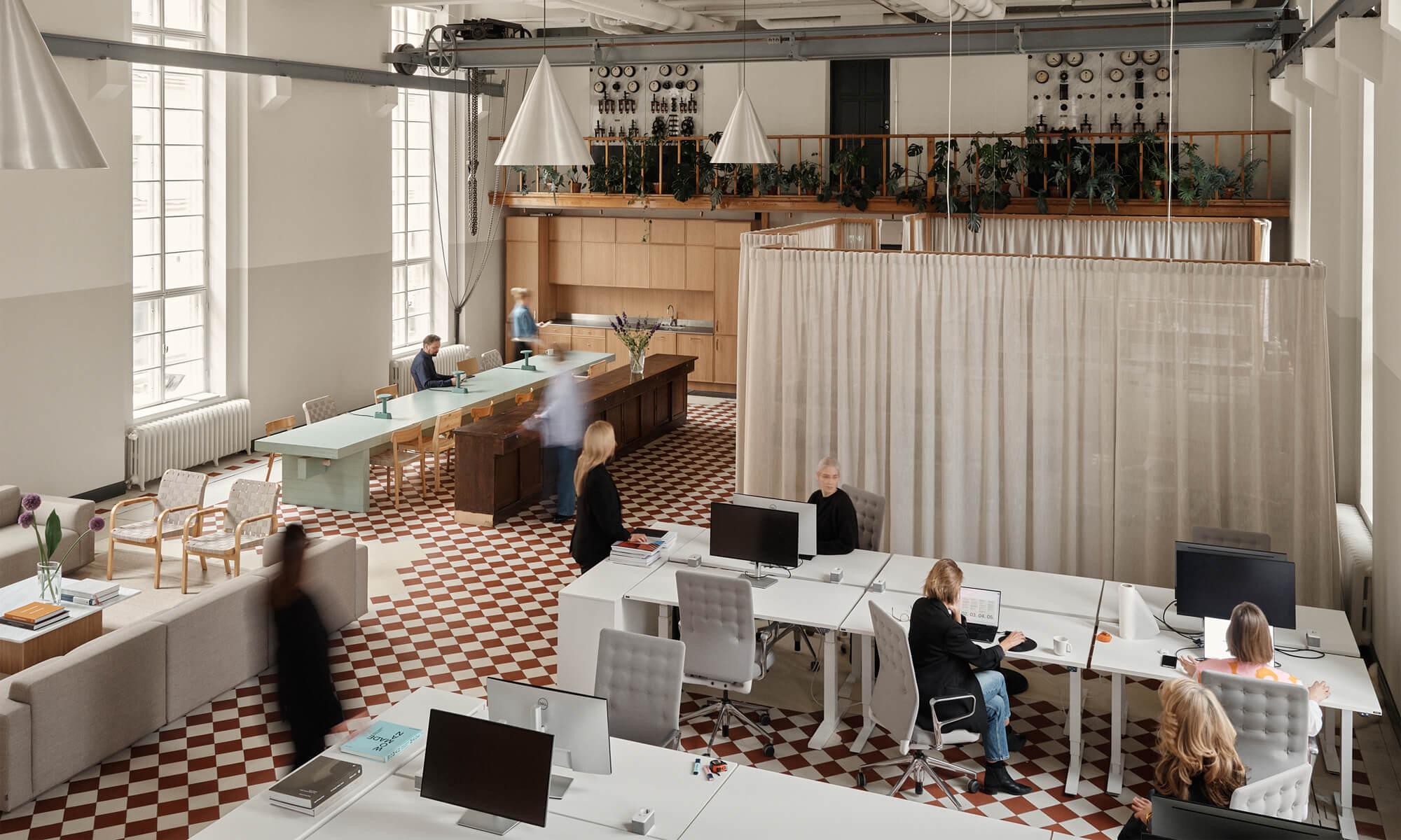 Spacious office with checkered flooring, large windows, and plants. Persons work at desks using computers in an organized, bright environment.