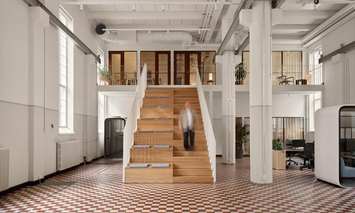 Spacious, modern office interior with a person descending wooden stairs. Features open workspaces, Framery smart pods, checkered floor, and white walls with large windows.
