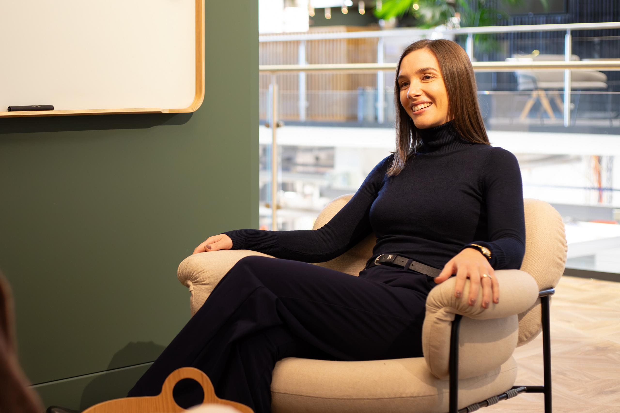 A person sits comfortably in a modern office chair, smiling, against a green wall with a whiteboard in a well-lit, spacious interior.