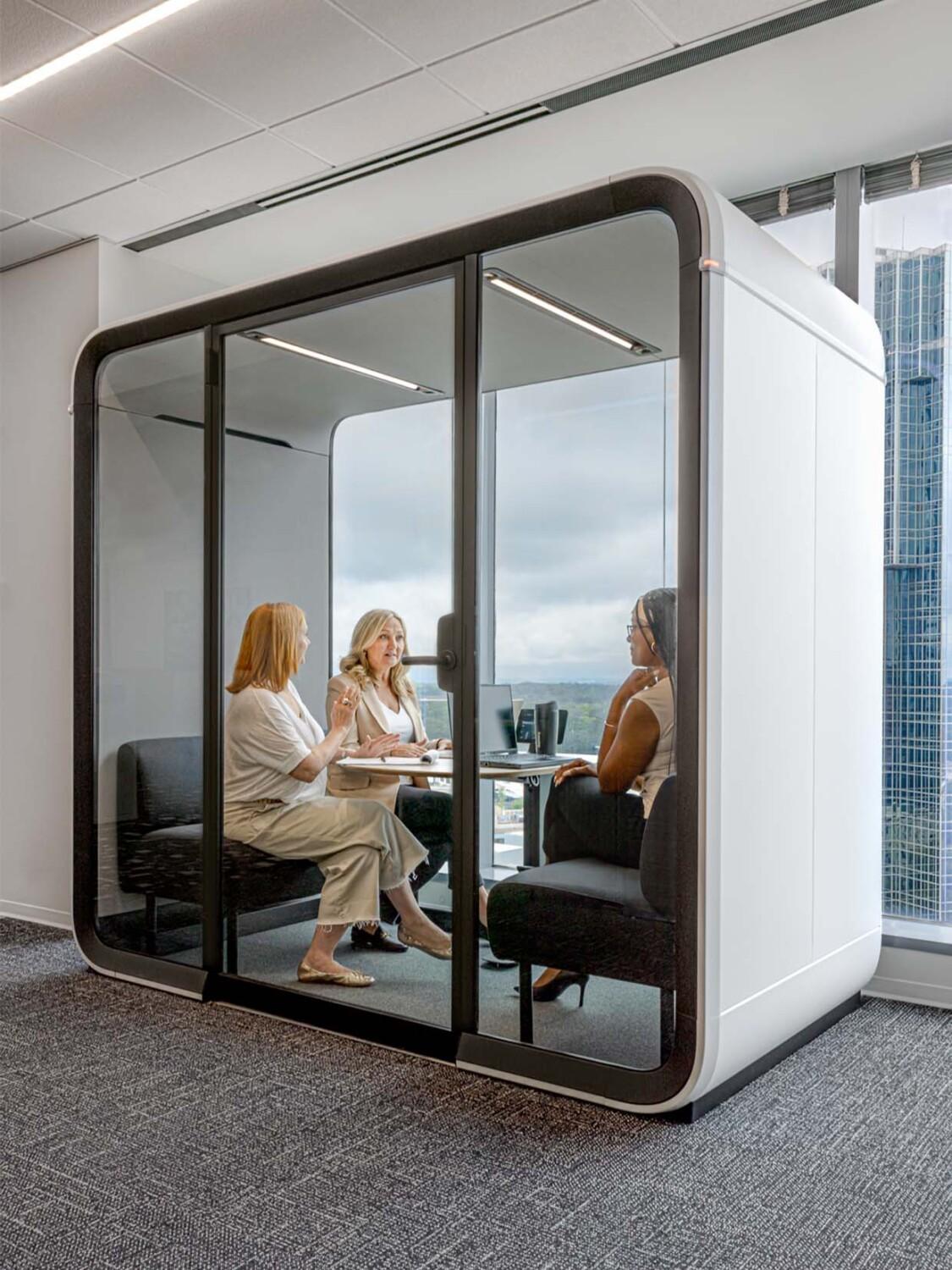 Three persons are in a modern glass meeting pod, situated in an office with cityscape views through large windows.