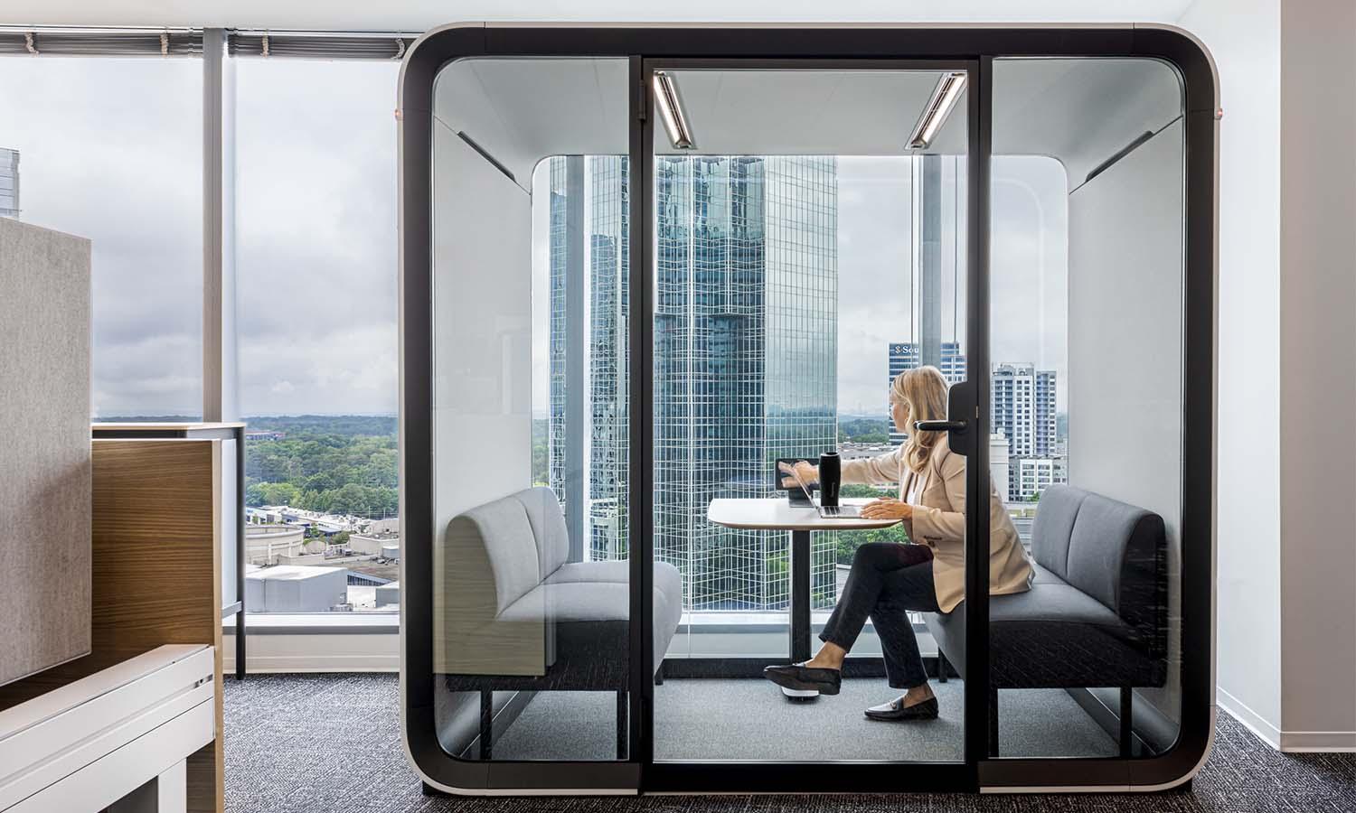 A person working in a Framery Four meeting pod next to a large window with an urban scenery.
