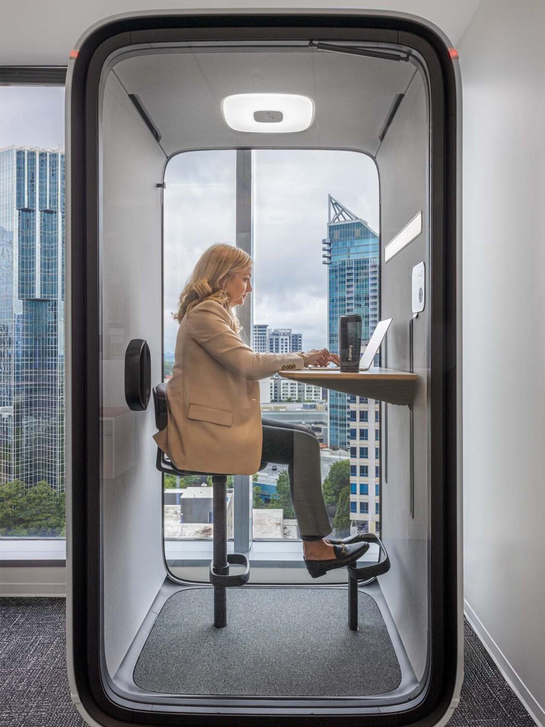 A person works on a laptop inside a modern office pod, with an urban skyline visible through the glass front.