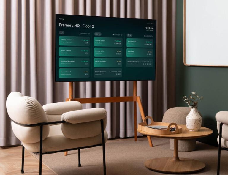 Modern office lounge with soft chairs, a wooden table, decorative vase, and a digital screen displaying room bookings at Framery HQ.