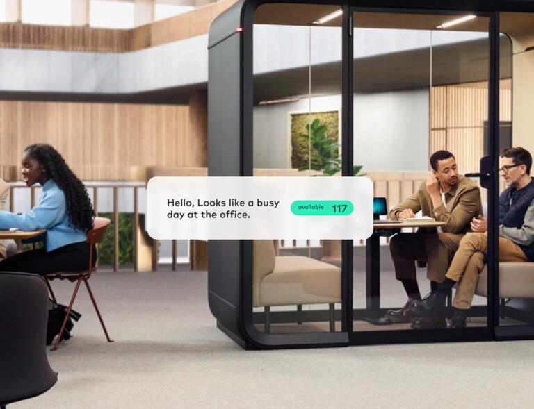 A modern office scene with two persons talking inside a Framery smart pod and others working outside. Digital display reads, "Hello, busy day."