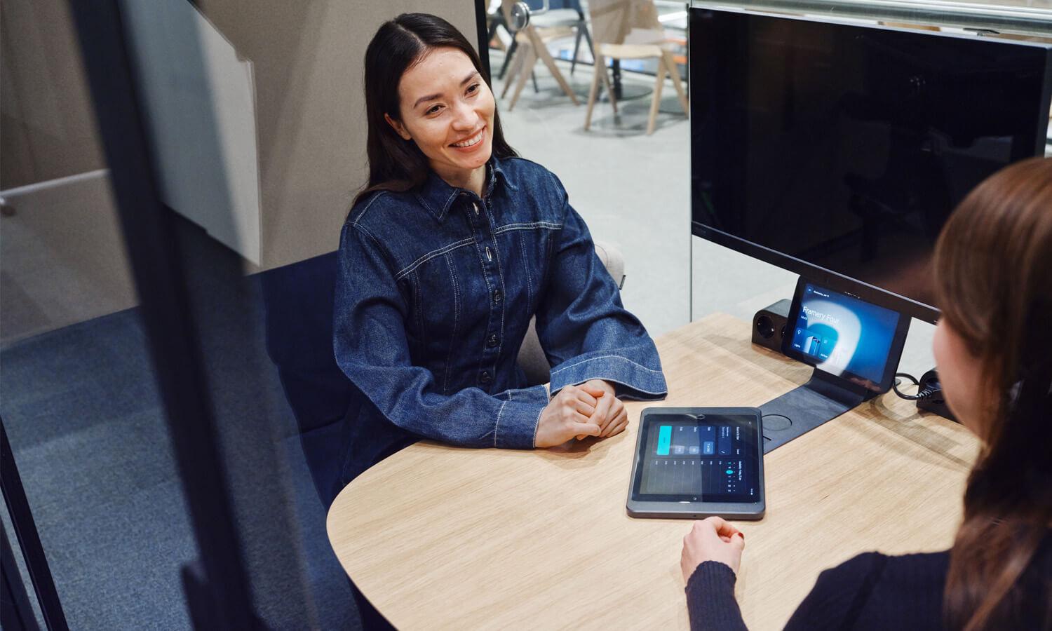 Two people sitting across a table in a modern office setting, engaged in conversation. Tablets and monitors with smart office solutions are visible on the desk.