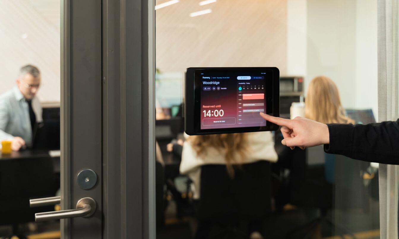 A tablet outside a glass-walled meeting room shows a 14:00 reservation; three people are seen working inside the room.