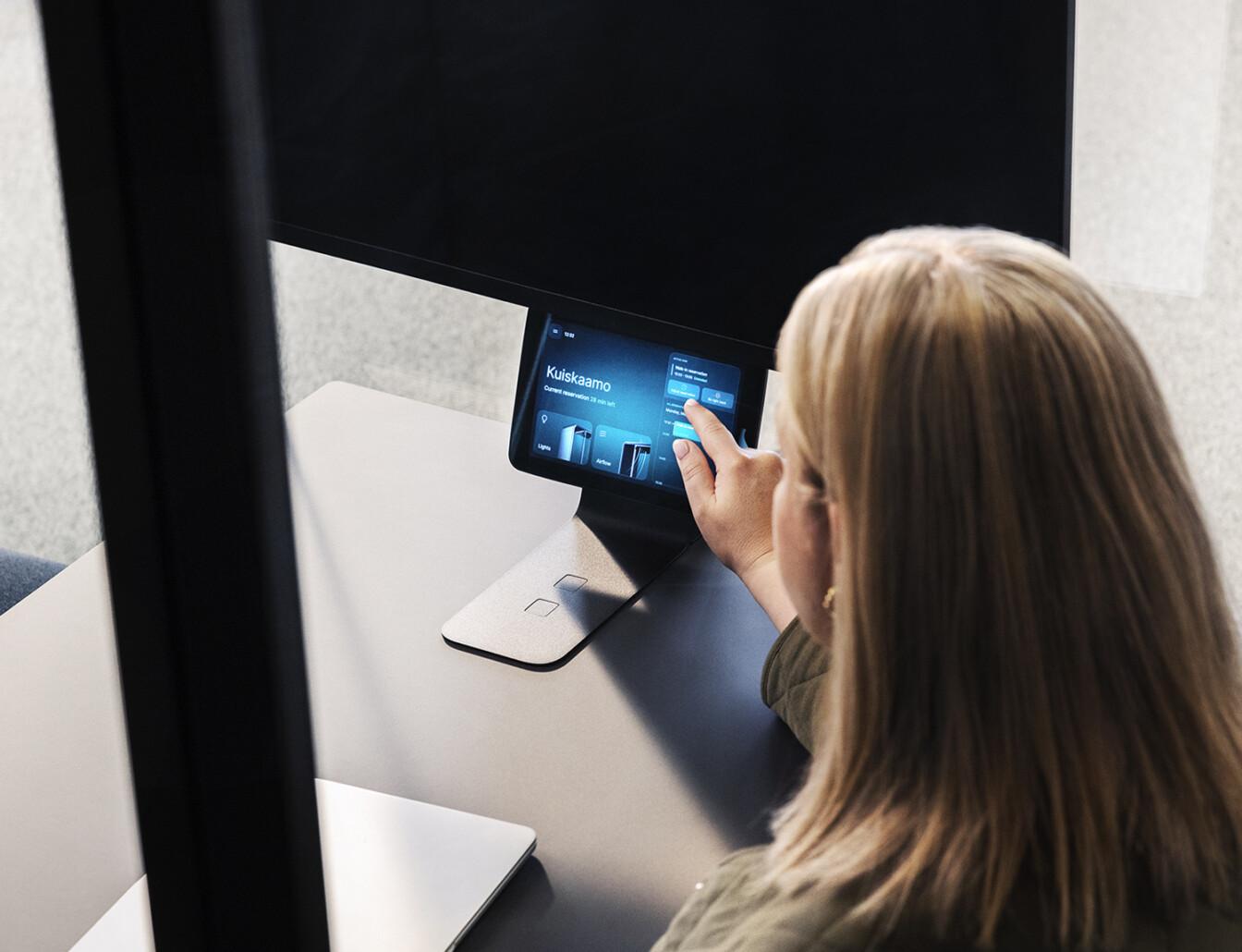 A person interacts with a touchscreen device displaying settings inside a modern, private office booth, focusing on energy and lighting adjustments.