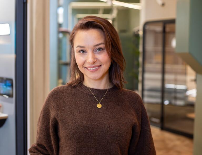 A person with brown hair smiles indoors, wearing a brown sweater and necklace. The modern interior features glass doors and neutral colors.