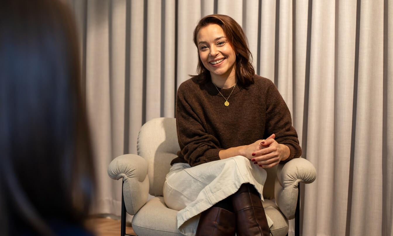 A person with brown hair sits in a chair, smiling, in a warmly lit room with curtains, talking to someone unseen.