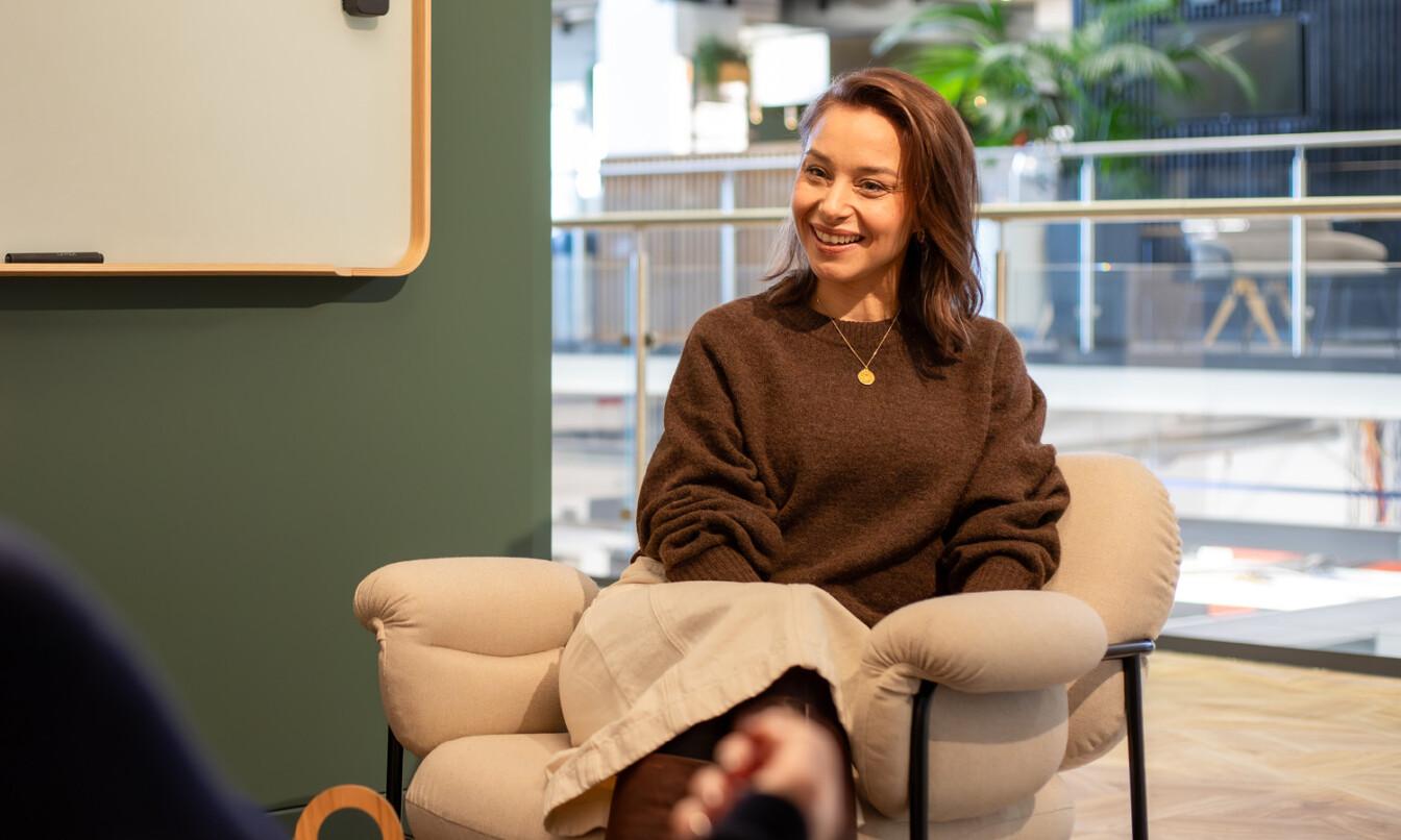 A person sits comfortably in a modern office setting, smiling, with a whiteboard on the left and plants in the background.