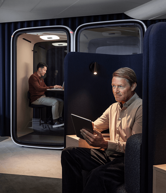 Two people working individually in modern, enclosed pods with tablets and laptops, seated in a professional, well-lit environment.